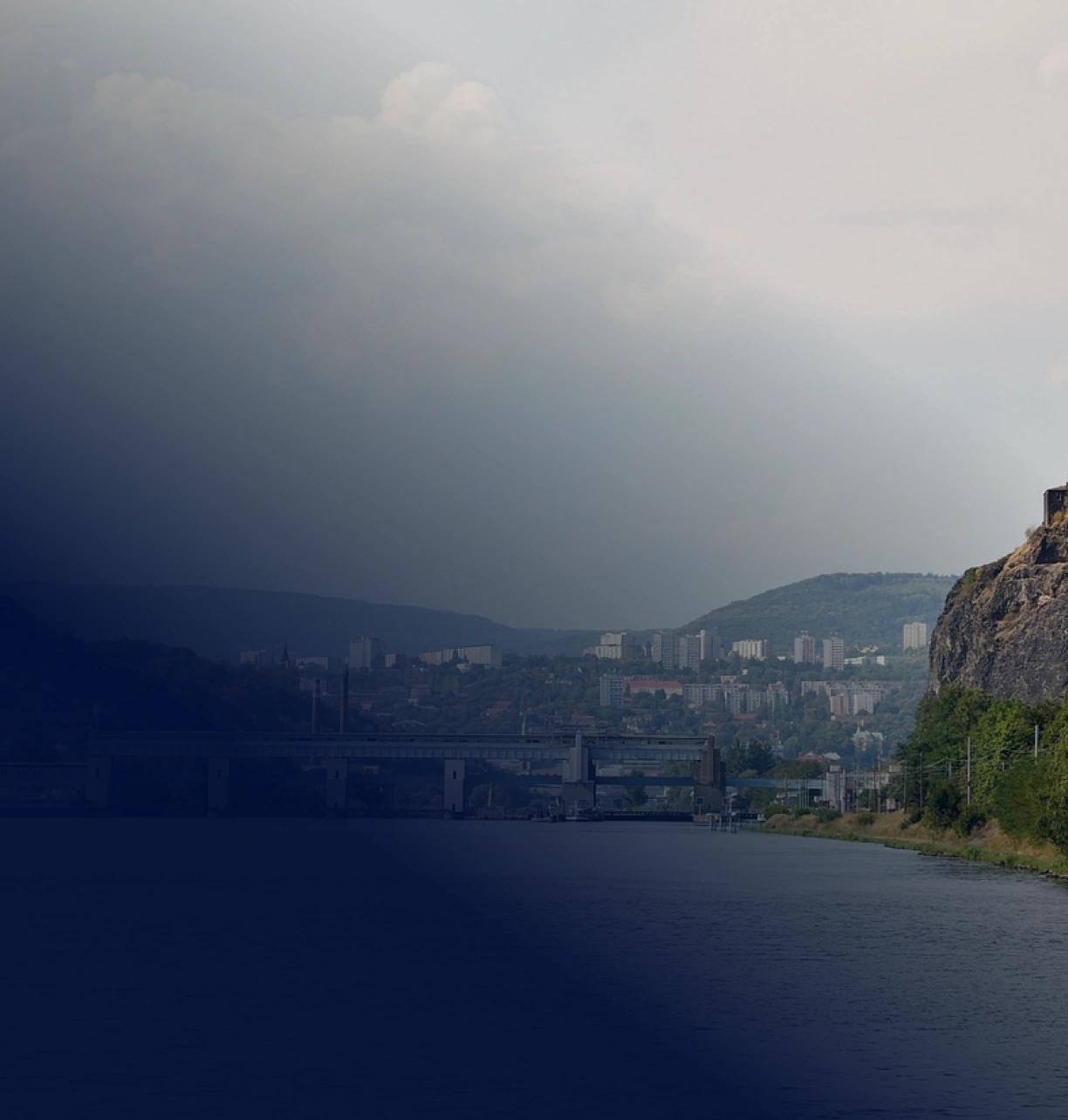 Pohled na Ústí nad Labem přes řeku, s plavebním zdymadlem, skalním svahem a městem v údolí pod zataženou oblohou