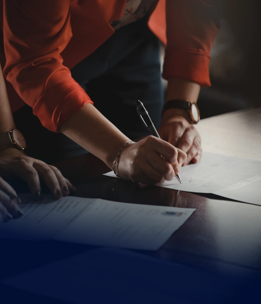 Osoba podepisuje dokument na stole, zatímco druhá osoba stojí vedle