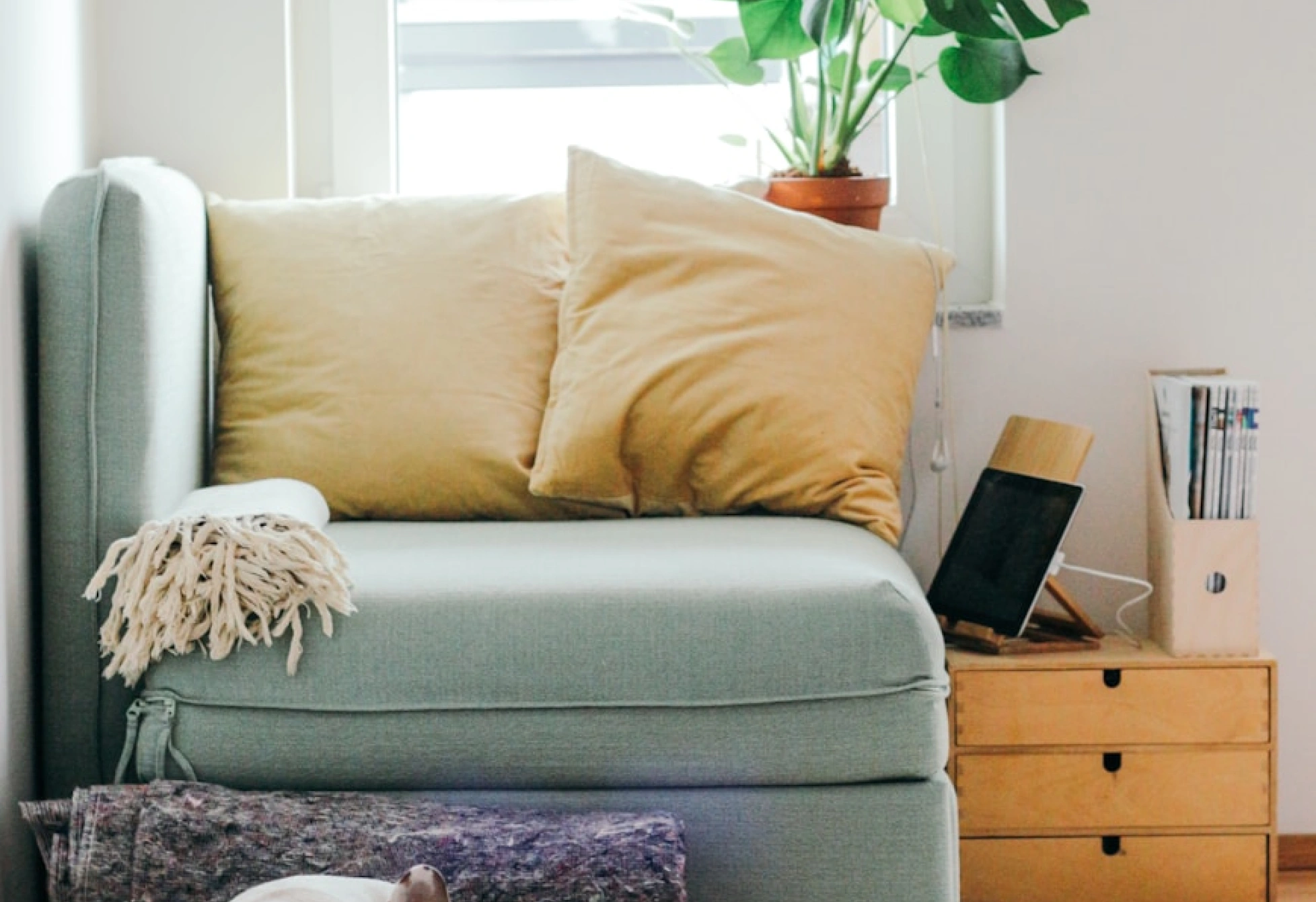 Detail světlého křesla nebo malé pohovky s dvěma béžovými polštáři, dekou na područce a dřevěným zásuvkovým stolkem vedle okna