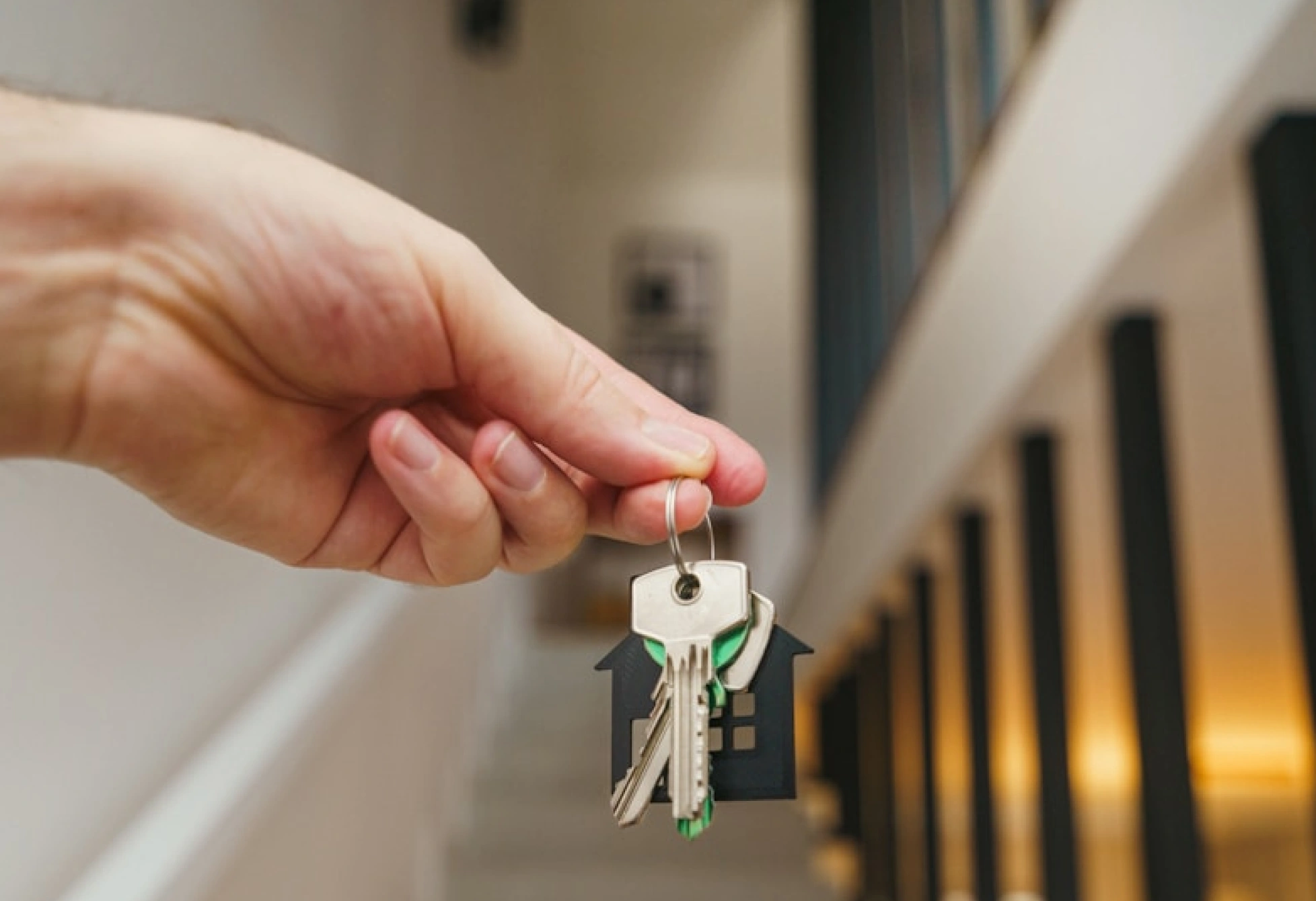 Ruka drží svazek klíčů s přívěskem ve tvaru domu na rozostřeném světlém pozadí
